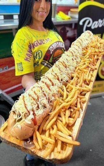 A smiling woman holds an extremely long hot dog, generously covered with melted cheese and sauce, served alongside a massive portion of french fries on a wooden board. Her yellow t-shirt has text, including 'HAMBURGUESAS' and 'JONRONER'.