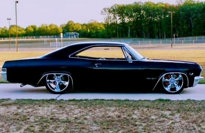 A black classic car with shiny chrome wheels parked on a dirt/grass field.