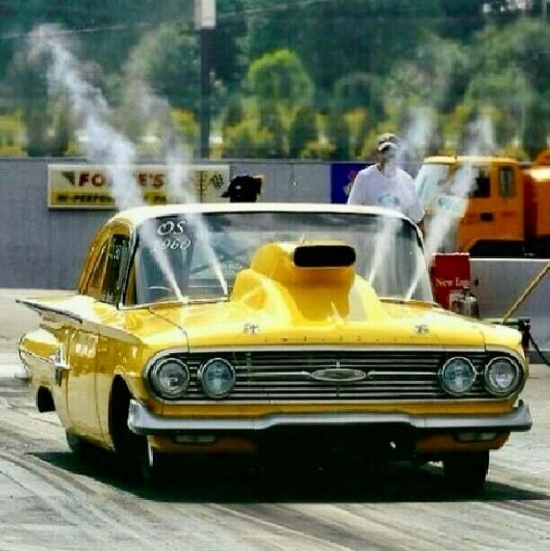 Yellow classic drag car with a large blower hood at a race track.