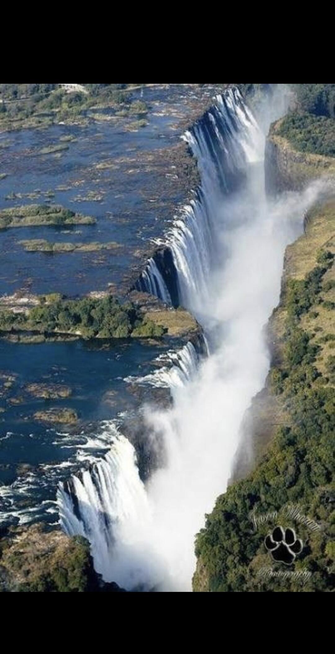 An aerial view of a majestic waterfall, possibly Victoria Falls, showing a vast curtain of water plunging into a deep gorge, surrounded by lush green landscape and a river upstream. A paw print logo is visible in the bottom right corner.