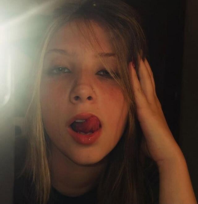 A young woman with her tongue out, looking at the camera. There's a bright light source on the left.