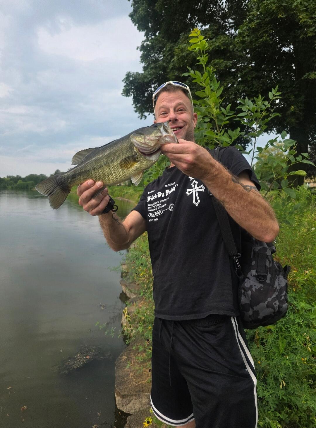 A man proudly holds a large bass fish by the mouth, smiling at the camera. He is wearing a black t-shirt with text 'BLESSED', 'FIGHT MY FAITH', 'GOD IS MY TOUGH BATTLE', 'STRONG SOLDIER', 'BEST HERE' and a cross symbol. He is standing by a river or lake with green trees and bushes in the background under an overcast sky.