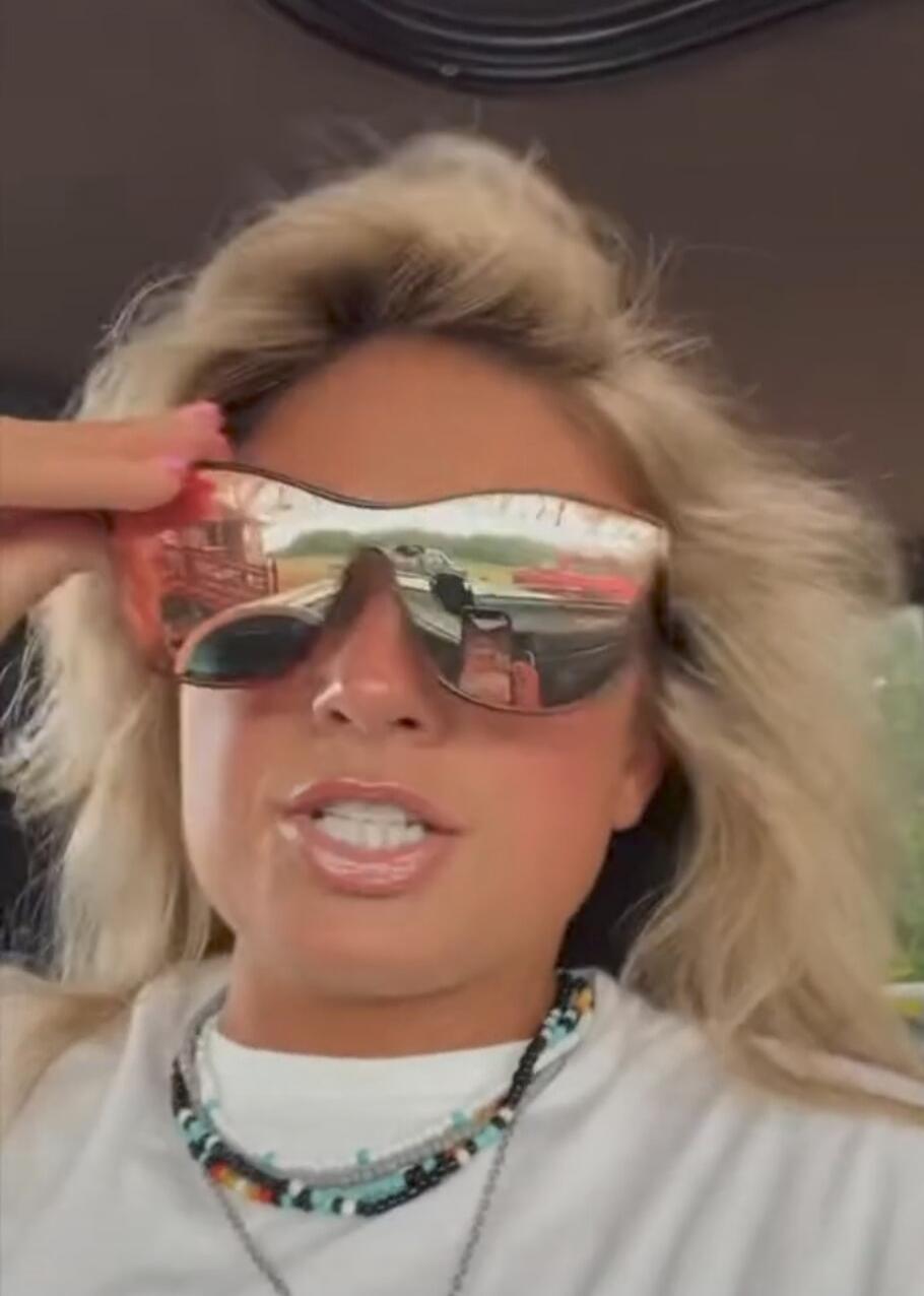 A woman is speaking while wearing sunglasses and beaded necklaces. She holds her sunglasses with one hand.