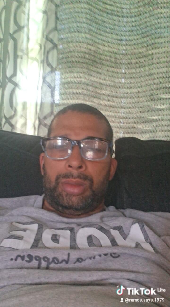 TikTok Lite @ramos.says.1979. A man wearing a grey t-shirt with 'MODE' (reversed) and other text (reversed) is lying on a couch.
