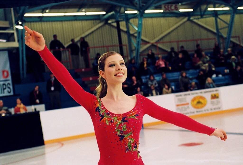 The image shows a young woman figure skater in a red costume with colorful embellishments, performing on an ice rink. She is smiling and looking upwards, with her left arm extended. The background shows spectators in the stands and the structure of an indoor arena.