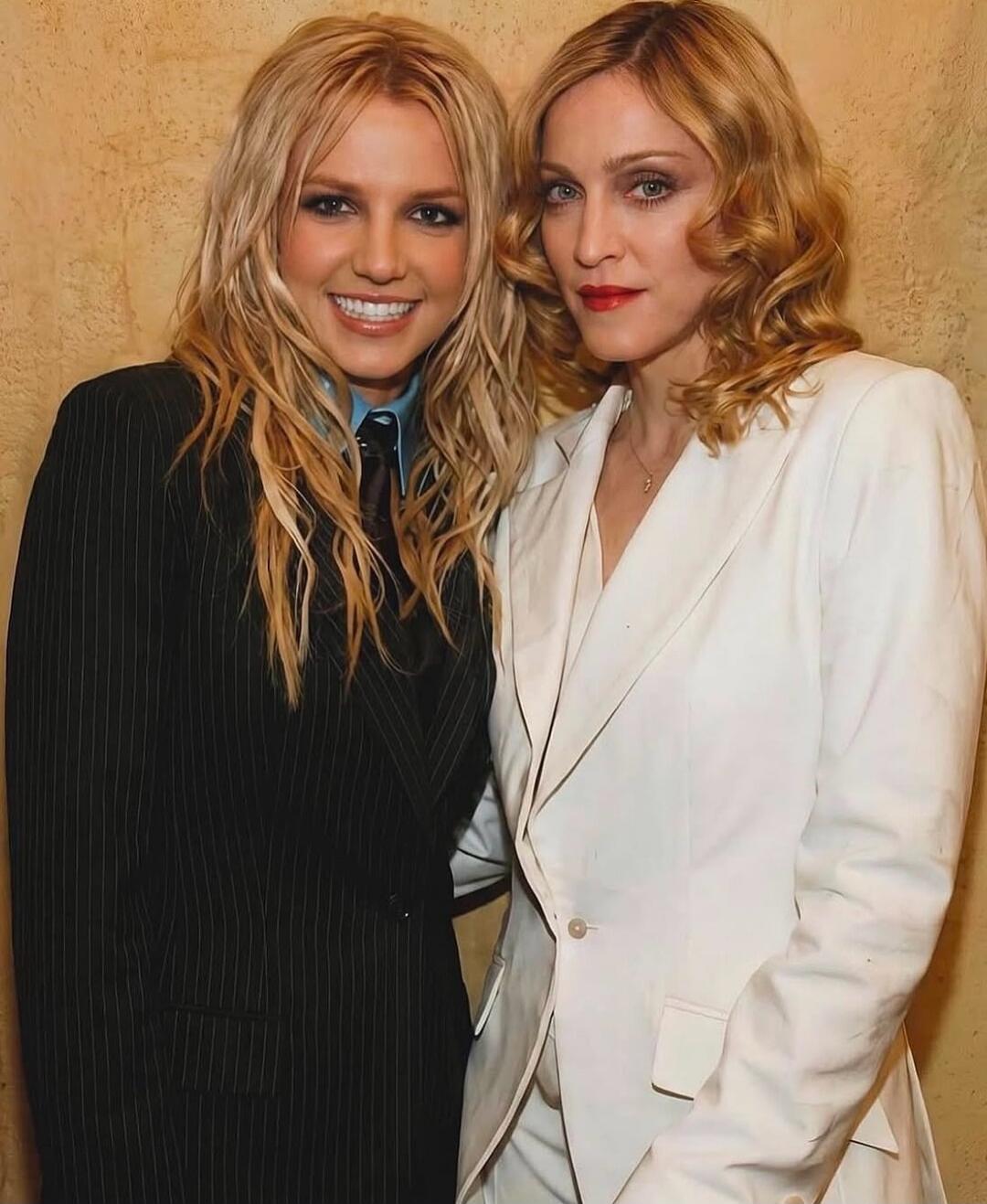 Two women posing for a photo, one in a black pinstripe suit, the other in a white suit.