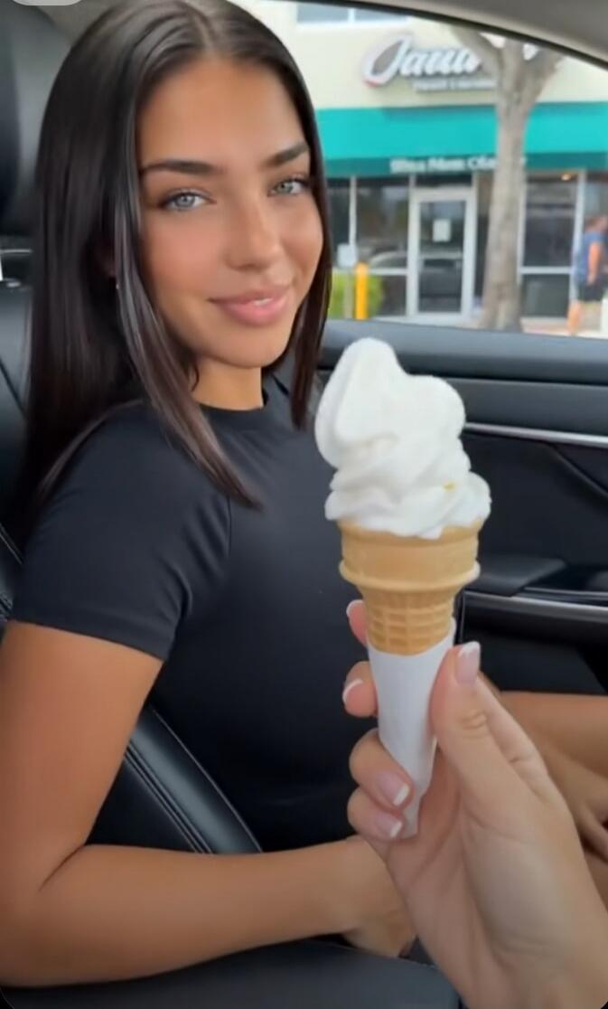 A woman in a car is offered a vanilla soft-serve ice cream cone. She smiles at the ice cream. In the background, a store sign reads 'Jawa'.