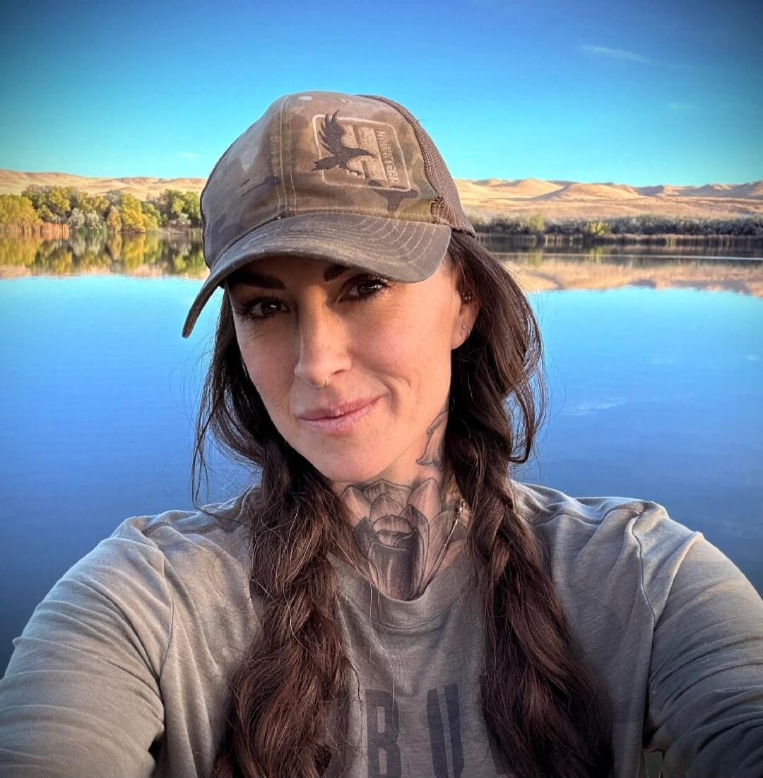 A woman wearing a camouflage cap with an eagle logo and 'NORTHWEST' text, and a t-shirt with partially visible text, possibly 'BU'. She has long braided hair and visible neck tattoos. She is taking a selfie outdoors by a calm body of water that reflects the blue sky and distant hills with some trees.