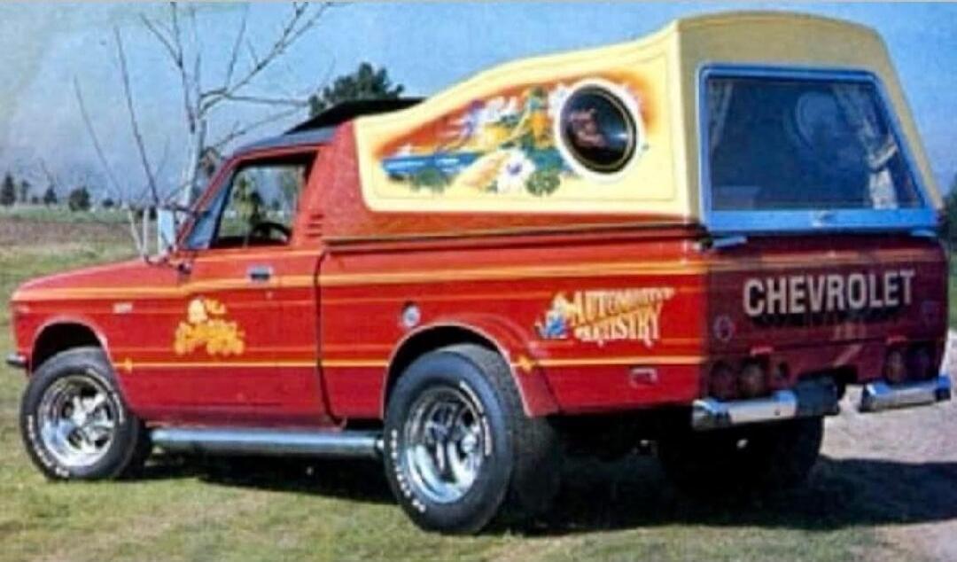 CHEVROLET. AUTOMOTIVE ARTISTRY. A vintage red Chevrolet pickup truck with a custom yellow camper shell featuring airbrushed artwork of a landscape scene. The truck also has custom decals and chrome side pipes.