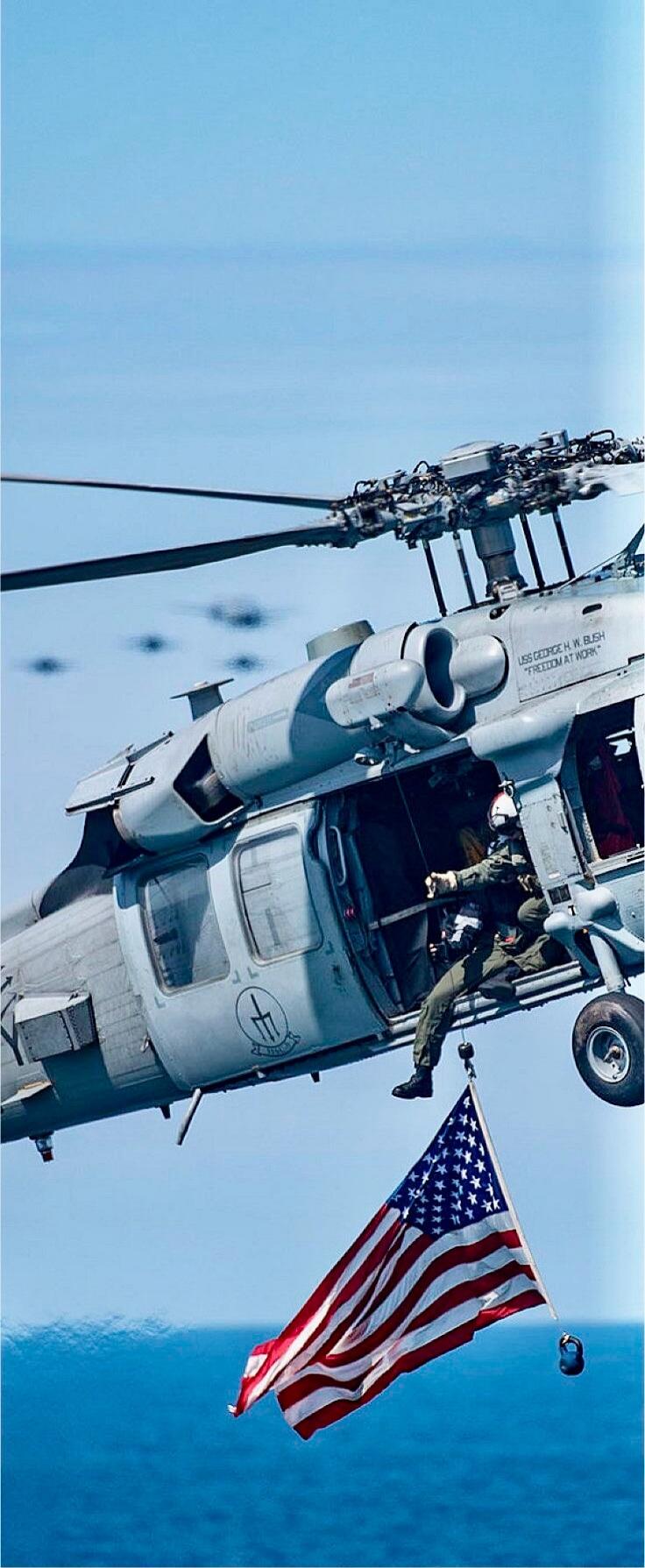 A military helicopter, identified as USS George H. W. Bush, with the motto 'Freedom at Work', flies over the ocean. A person is seen hanging from the side, holding a large American flag.