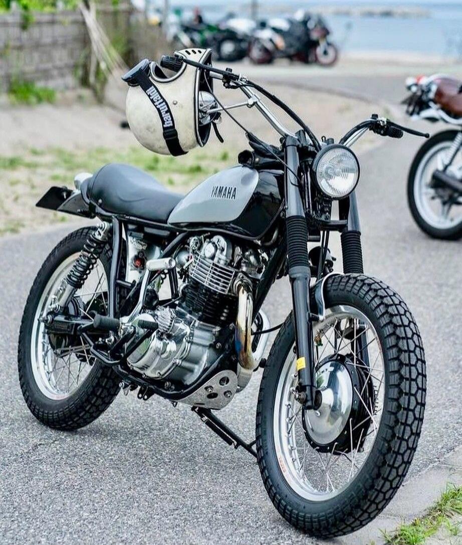 A vintage Yamaha motorcycle parked on a street, with a helmet hanging from the handlebar. Background shows a coastal area with other bikes in the distance.