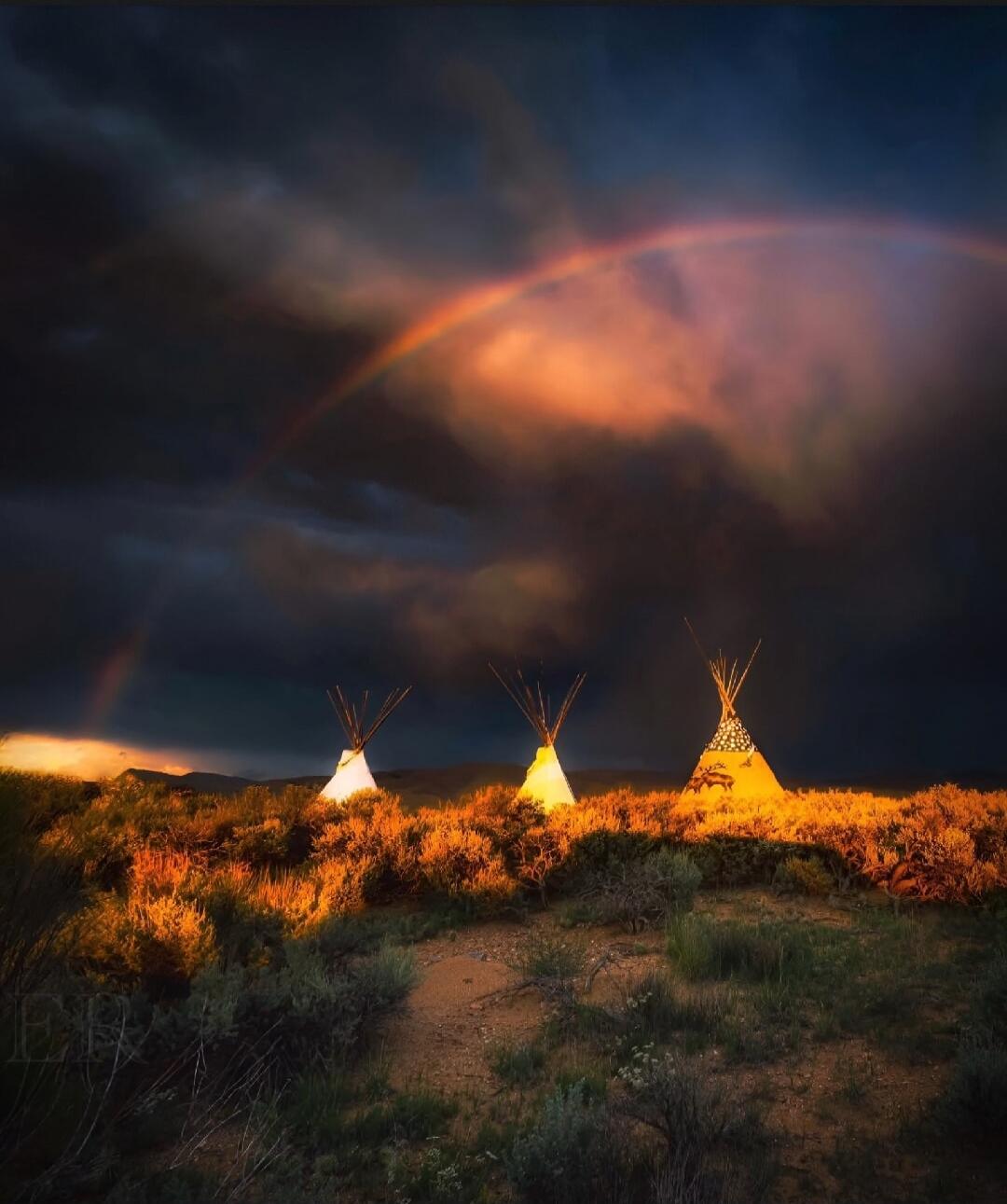 Three tipis on a desert plain at sunset with a rainbow in the cloudy sky.
