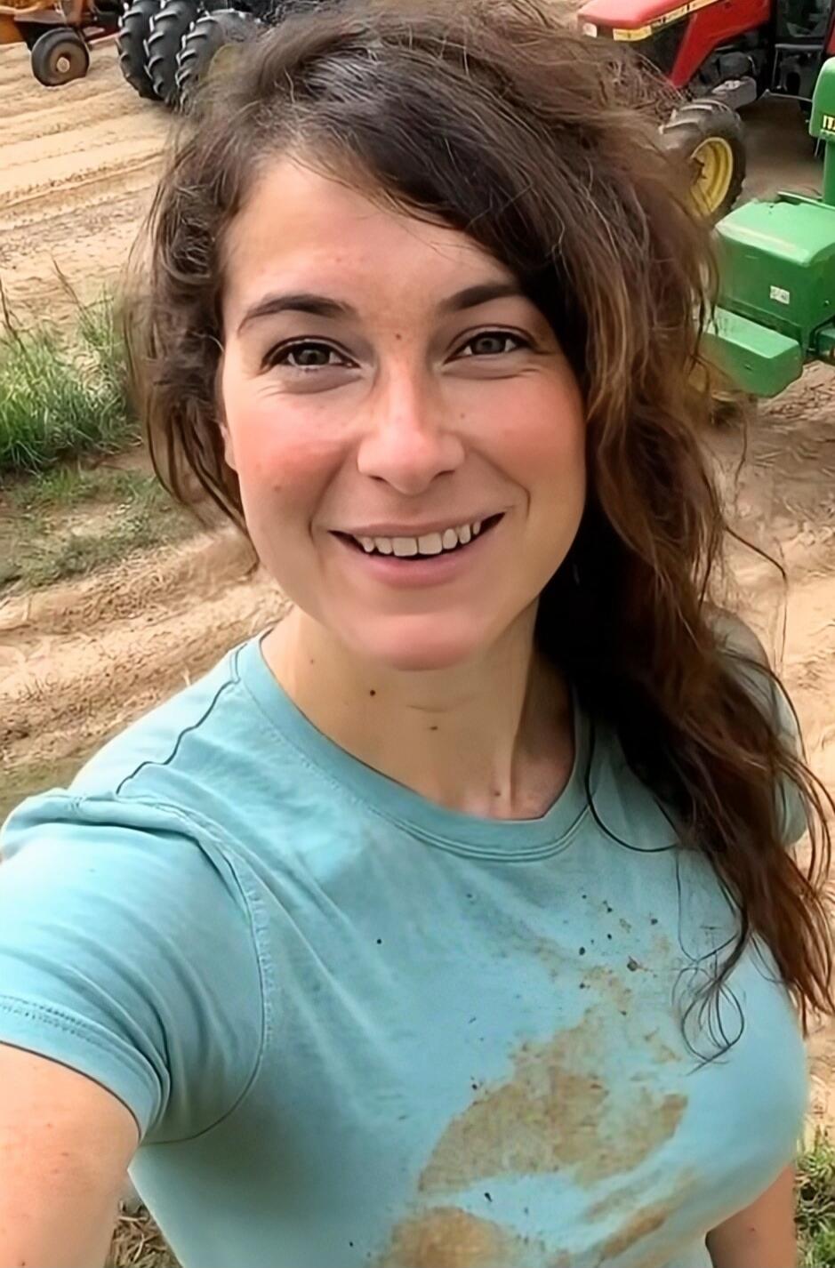 A woman taking a selfie outdoors on a dirt field, with dirt on her light blue shirt.