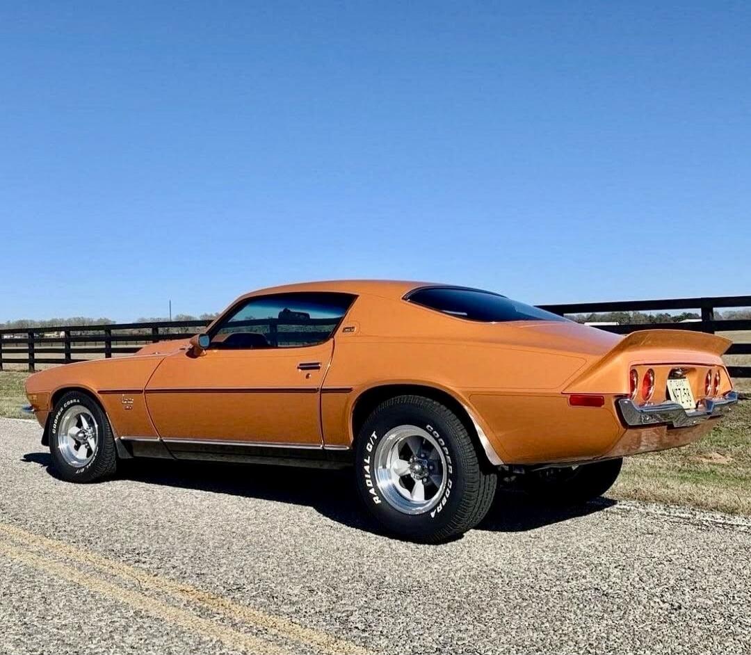 A classic orange Chevrolet Camaro SS with Cooper Cobra Radial G/T tires, license plate NET 543, parked on an asphalt road next to a wooden fence under a clear blue sky.