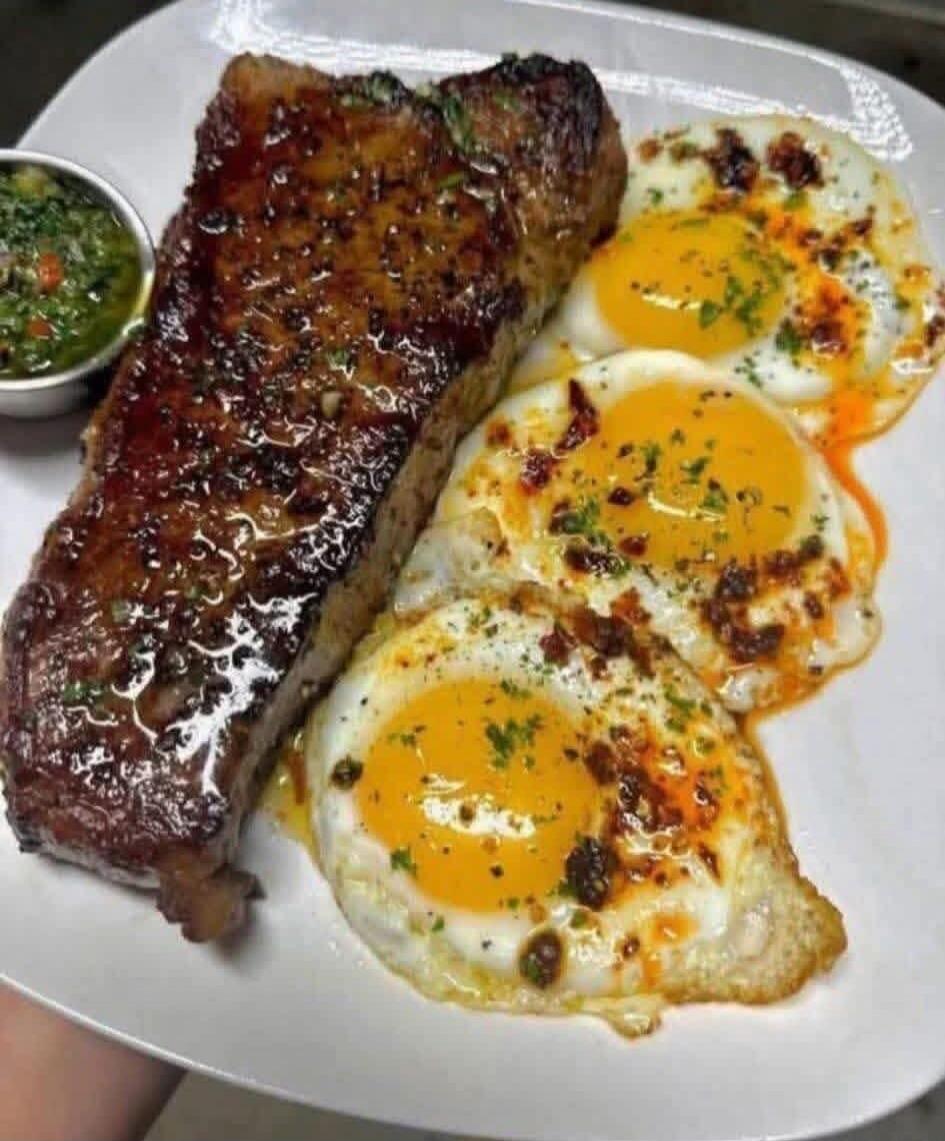 A plate with a large glazed steak and sunny-side-up eggs, garnished with herbs. A small cup of green sauce sits on the side.