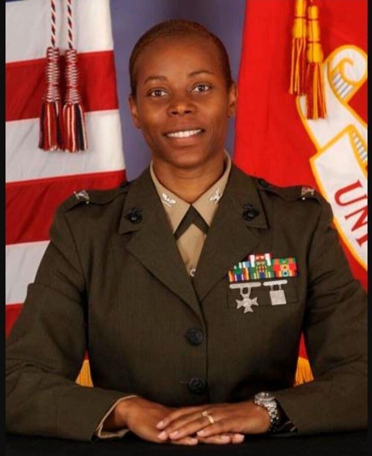 A woman in a military uniform, possibly a US Marine, is smiling and looking at the camera. She is wearing medals and ribbons. Flags are visible in the background, including a partial 'UN' from 'US MARINE CORPS'.