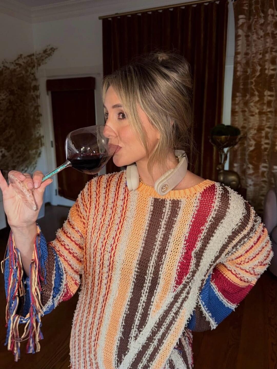 A woman in a colorful striped sweater is drinking red wine from a large glass. She has headphones around her neck and her hair is in a bun. The setting appears to be indoors.