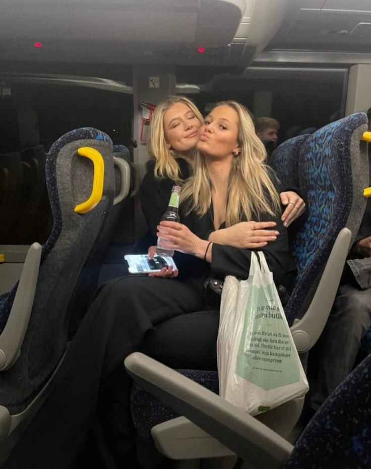 Two women sitting on a train, kissing. One holds a bottle and a phone; there is a white bag on her lap.