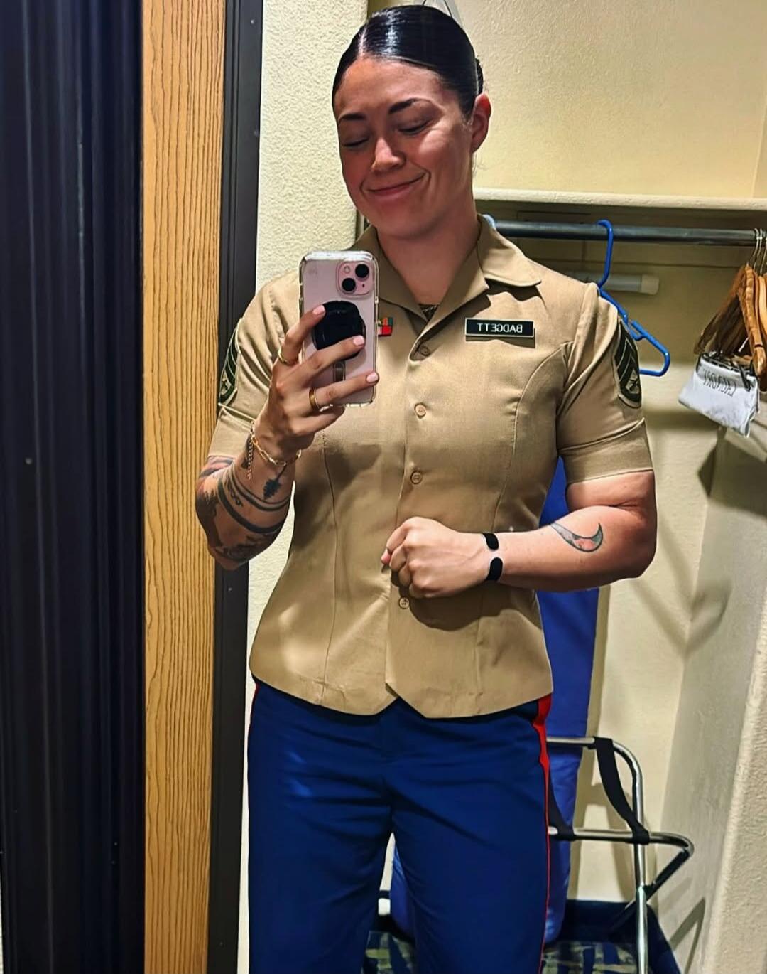 A woman in a military uniform taking a mirror selfie. She is smiling with her eyes closed. Her uniform shirt is khaki, and her pants are blue with a red stripe. She has tattoos on her arms. The name tag on her uniform reads 'BADGELL'.
