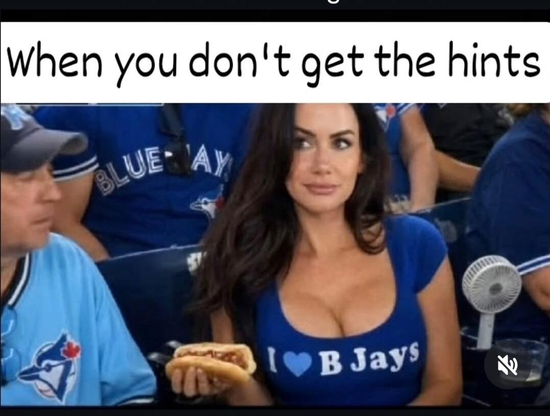 when you don't get the hints. A woman in a low-cut blue shirt with 'I ❤️ B Jays' printed on it, holding a hot dog, looks at a man who is looking away. The text on her shirt is a double entendre, referring to both the Toronto Blue Jays baseball team and her breasts.
