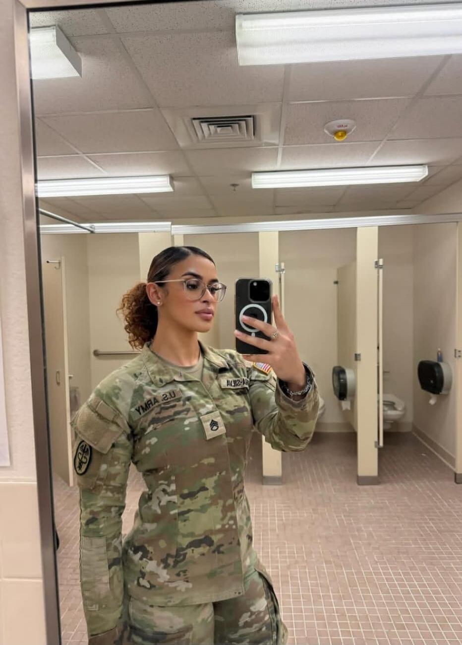 A woman in a US Army uniform (ACU/OCP) takes a mirror selfie in a public restroom. She is wearing glasses and has her hair tied back. The uniform shows 'U.S. ARMY' (mirrored as YMRA 2.U), a Staff Sergeant rank insignia, and an American flag patch. She is holding a smartphone with a MagSafe ring.