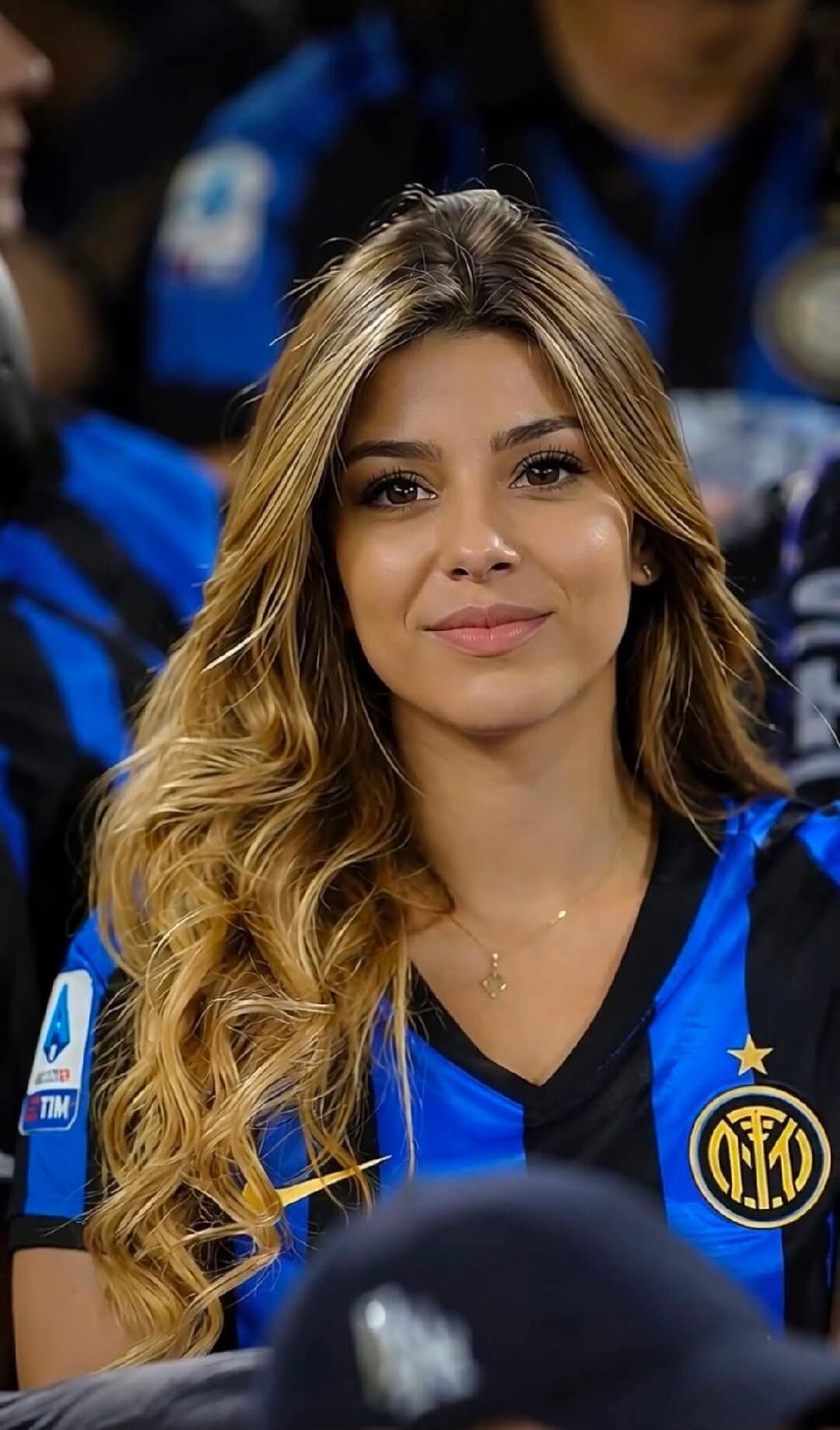 A young woman with long blonde hair wearing an Inter Milan football jersey, smiling at the camera in a stadium setting.