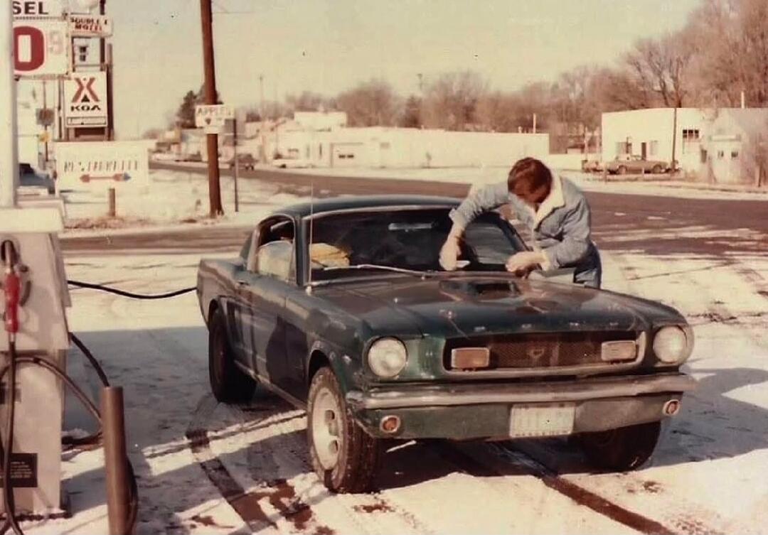 SEL 0.9 X KOA APPLE REST AREA. A man is cleaning the windshield of a classic dark green Ford Mustang at a gas station in a snowy, winter setting.