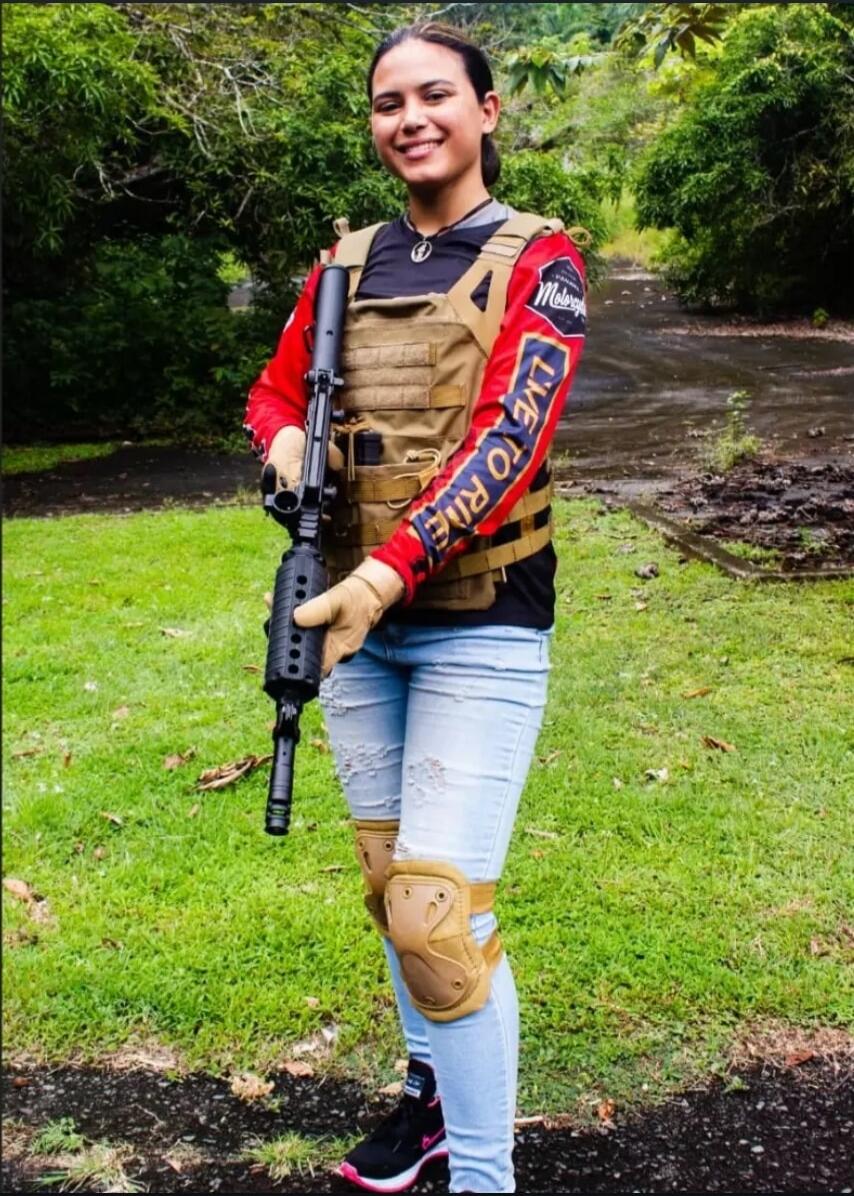 A young woman wearing a tactical vest, ripped jeans, knee pads, and a red long-sleeve shirt with 'LIVE TO RIDE' text, holding a rifle and smiling. The background is a green outdoor area with trees.