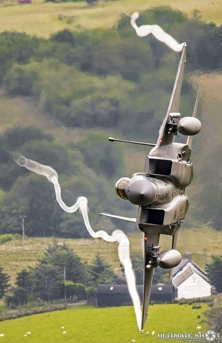 ULTIMATE SHOT. A military jet, possibly a Tornado, is captured in a dynamic low-altitude maneuver, leaving distinctive vapor trails from its wingtips. The aircraft is banking sharply against a backdrop of green hills, fields with sheep, and scattered rural buildings.