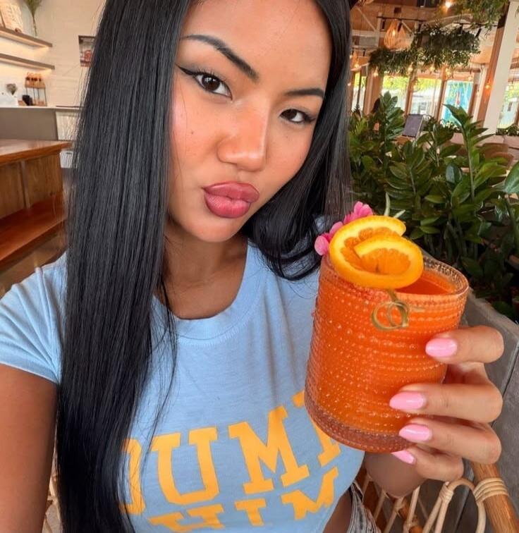 A woman wearing a light blue t-shirt with yellow text 'UM' is holding an orange drink garnished with orange slices and small pink flowers. She has long dark hair, full lips, and winged eyeliner. The background shows an indoor setting with plants, possibly a cafe or restaurant.