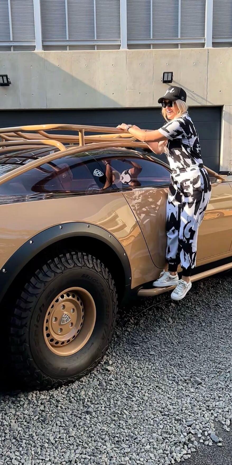 A woman in a black and white patterned jumpsuit, sunglasses, and a black cap with an 'H' logo is smiling while standing on the side step of a custom gold-colored car. The car features large off-road tires with 'BFGoodrich' visible and a 'Maybach' logo on the wheel hub and side window. She is holding onto the gold-colored roof rack. The setting is o