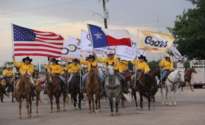 A group of people on horseback, dressed in yellow shirts and cowboy hats, participating in a parade or event. They are carrying various flags, including the American flag, the Texas state flag, and a flag with the 'Coors' logo. Other flags show symbols like a horseshoe and text that appears to be '20 years'.