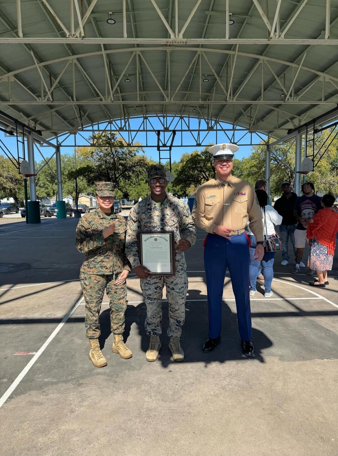Three Marines, two in camouflage utility uniforms and one in a dress uniform, are standing on a basketball court under a covered area. The Marine in the center is holding a framed certificate. All three are smiling. In the background, several civilians are visible. The certificate appears to be from the United States Marine Corps.