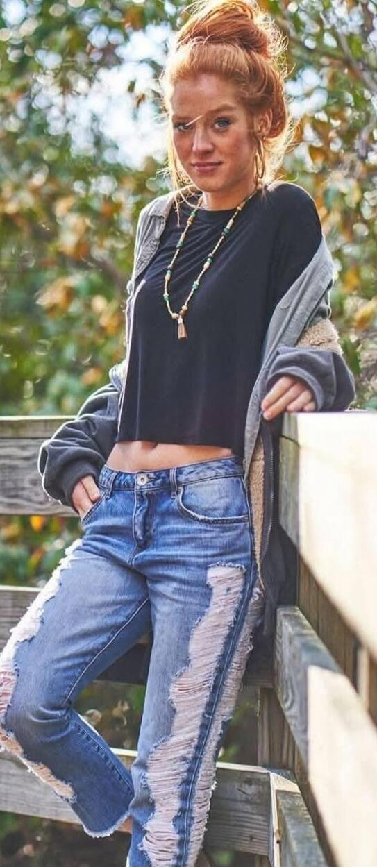 Young woman standing outdoors, leaning on a wooden railing. She is wearing a black cropped top, ripped jeans, a grey cardigan draped over her shoulders, and a beaded necklace.
