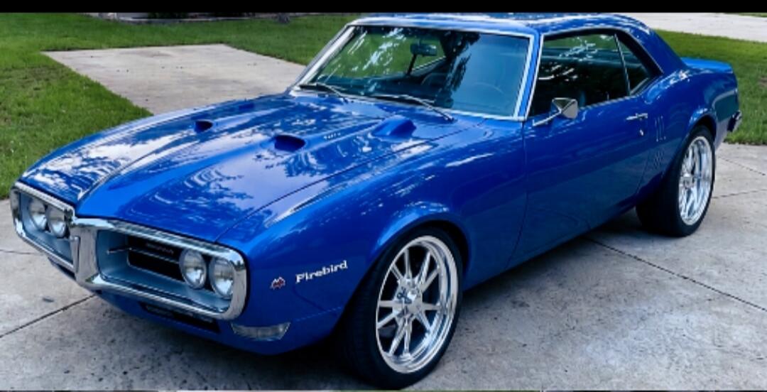 A classic blue Pontiac Firebird car, parked on a concrete driveway. The car has custom chrome wheels. The word 'Firebird' is visible on the side.