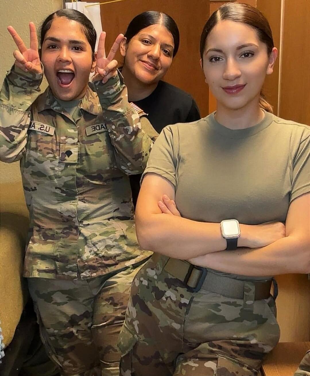 Three women, two in military camouflage uniforms, posing for a photo. One woman on the left makes peace signs and smiles widely, while another on the right crosses her arms and smiles subtly.