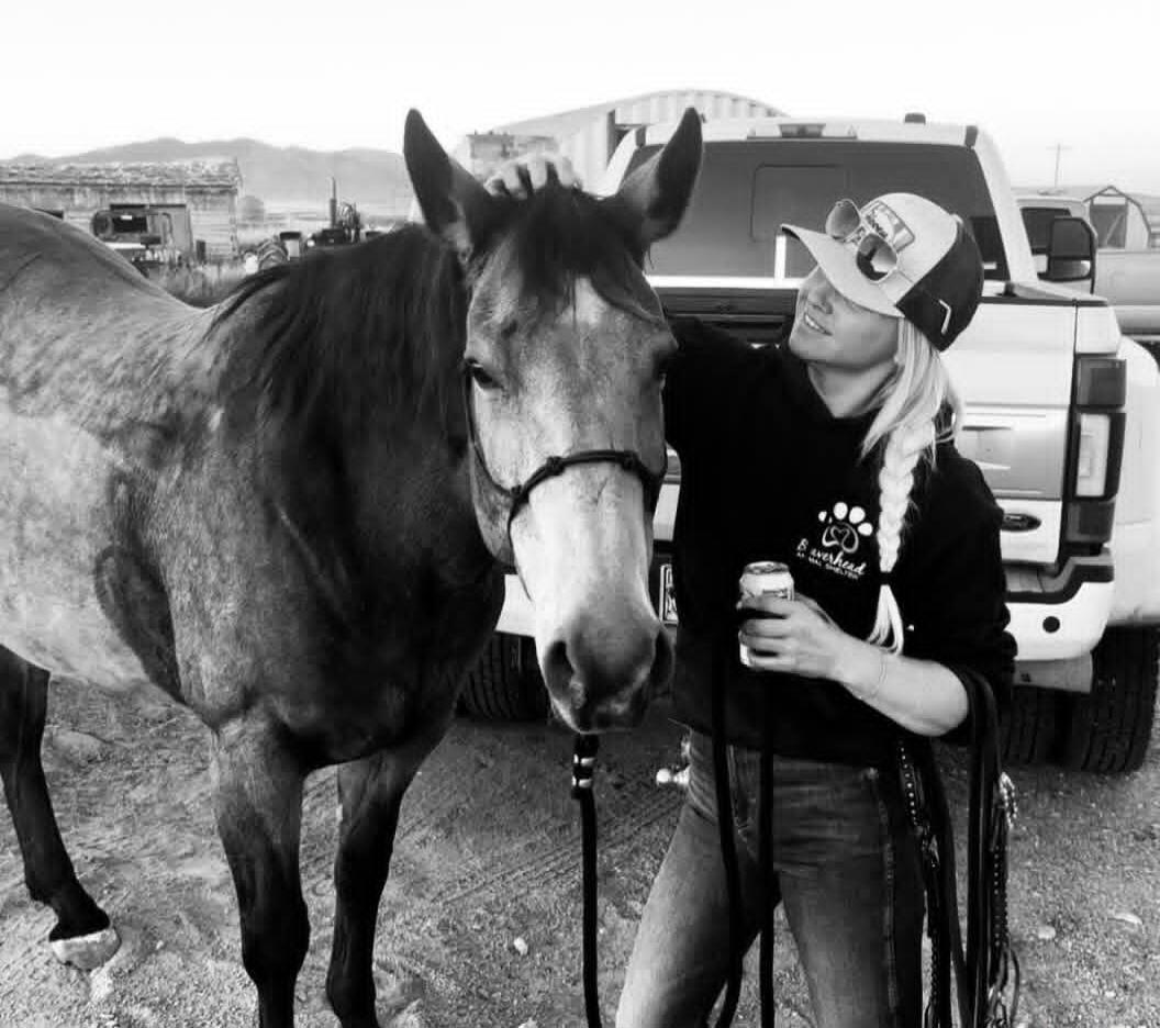 A woman with a braided blonde ponytail, wearing a cap and a hoodie with a paw print logo and text that might say 'overshead' or 'overshelter', is gently petting a horse. She holds a can in one hand and reins in the other. A white pickup truck is visible in the background, suggesting a rural or ranch setting.