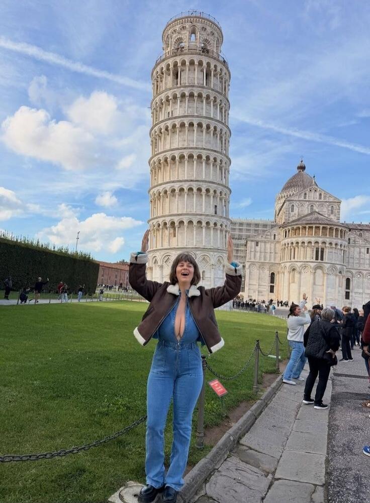 A woman is posing playfully in front of the Leaning Tower of Pisa, appearing to hold it up. She is wearing a denim jumpsuit with a very deep V-neck, revealing significant cleavage. The sky is blue with some clouds, and other tourists are visible in the background.