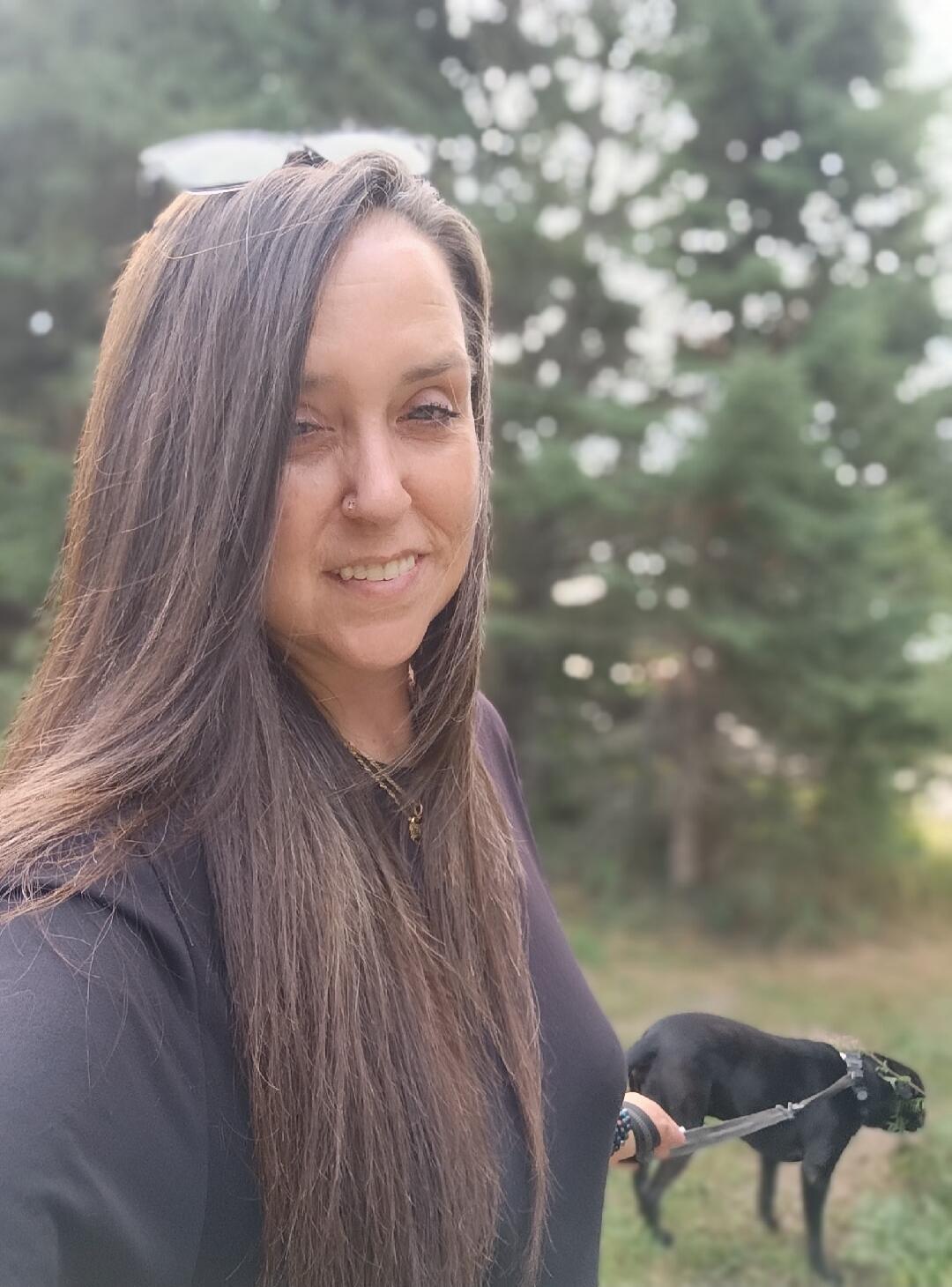 A woman with long brown hair smiles at the camera while holding a leash with a black dog in a green outdoor setting.