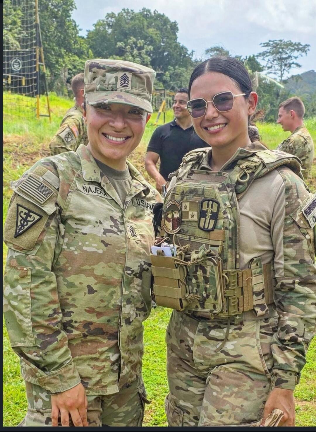 Two women in military camouflage uniforms are smiling at the camera. One has the name 'NAJERA' and 'U.S. ARMY' visible on her uniform, along with an American flag patch and a Master Sergeant rank insignia on her cap. The other woman wears a tactical vest with a 'FILIPINAS' patch and sunglasses. They are outdoors, with other people and trees in the 