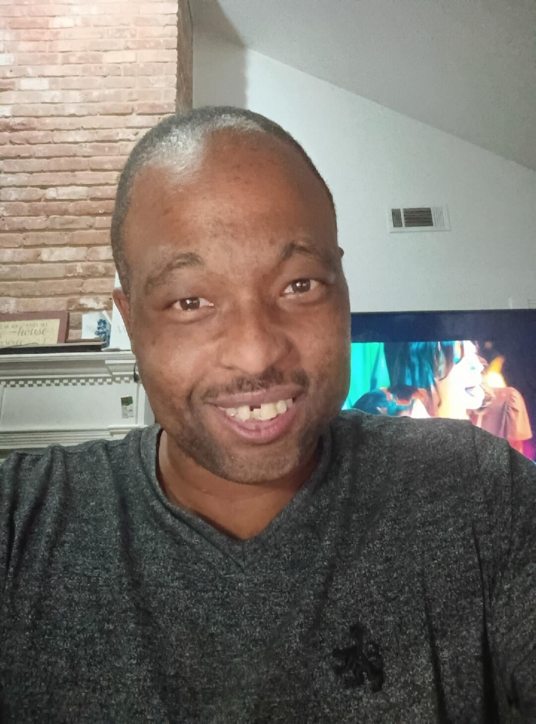 A man is smiling and taking a selfie. Behind him, a decorative sign reads 'THIS IS ME AND MY house'. A television screen is visible in the background.