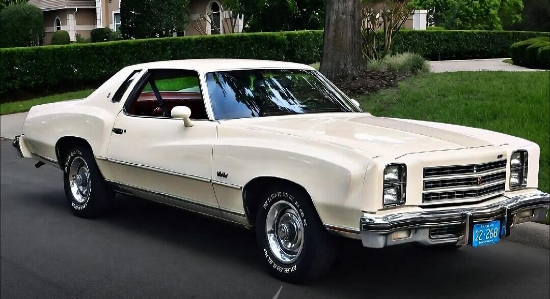 A vintage white Chevrolet Monte Carlo car is parked on an asphalt driveway next to a green lawn and hedges. A house is visible in the background. The car has chrome details and a blue Tennessee license plate showing '22-268' and 'VOLUNTEER STATE'.