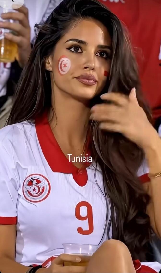 Tunisia 9. A woman with long dark hair, wearing a white and red sports jersey with the number 9. She has the Tunisian flag painted on her cheek and is holding a drink.
