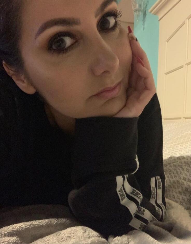 A woman with makeup and long eyelashes is lying down, looking at the camera. She is wearing a dark long-sleeved top with 'PINK' written on the sleeve. Her hand is resting on her chin.