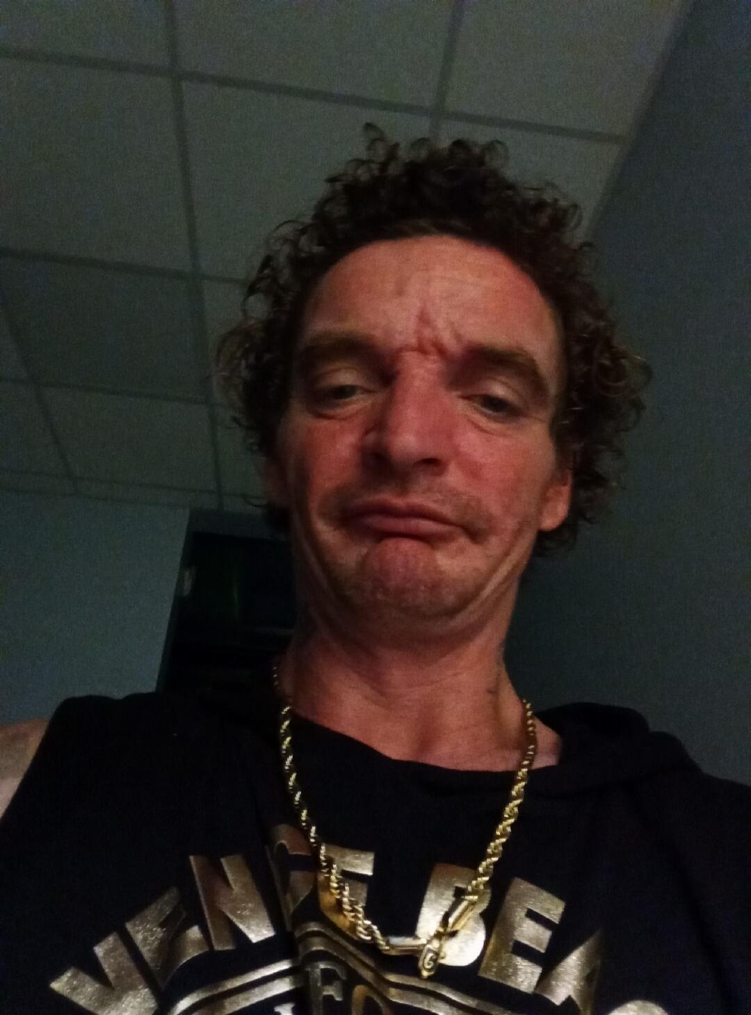 A man with curly hair and a gold chain takes a selfie, making a grumpy or confused face. His black tank top has 'VENICE BEACH' written on it.