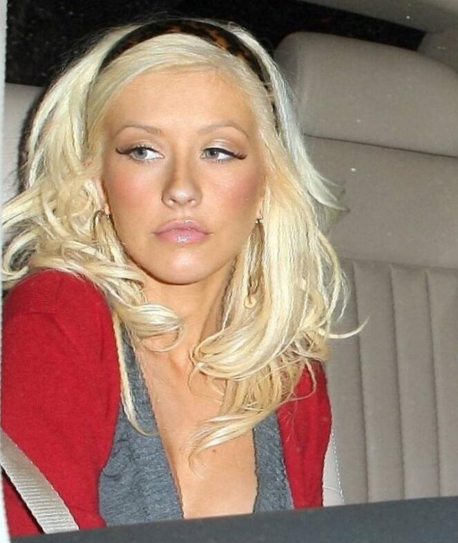 A blonde woman with wavy hair sits in a car, wearing a red cardigan over a gray top.