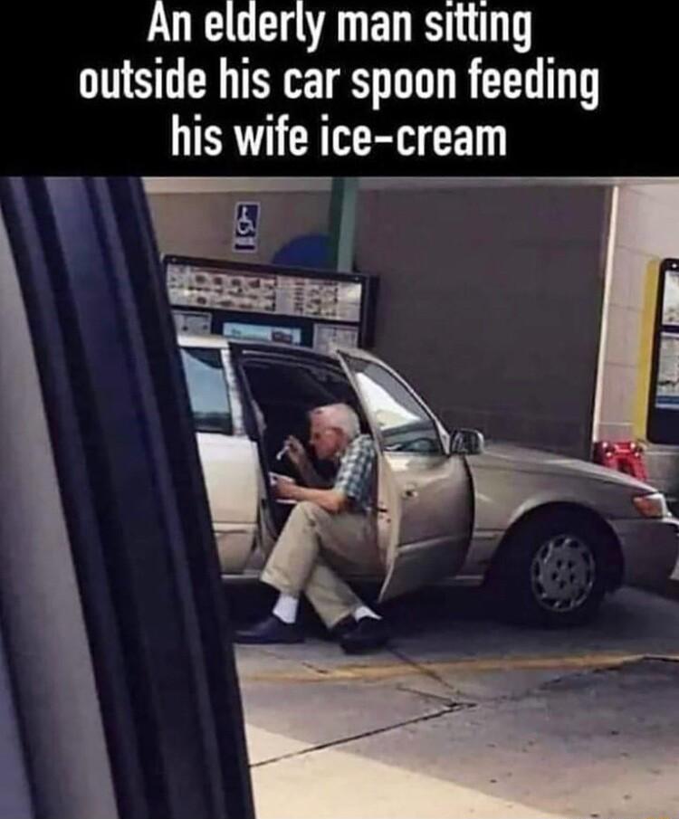 An elderly man sitting outside his car spoon feeding his wife i ice cream