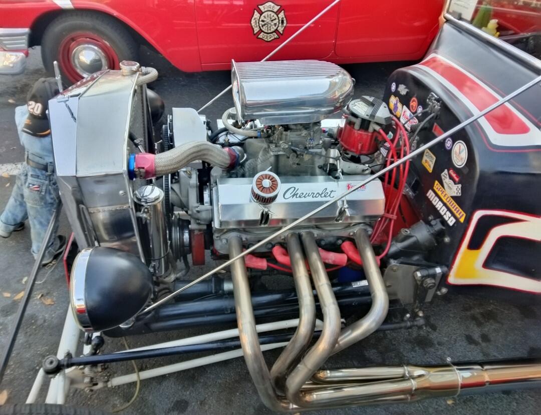 A custom Chevrolet engine is prominently displayed in a hot rod, featuring chrome components and performance stickers like 'SPEEDWAY' and 'MOROSO'. In the background, a red classic car with a 'FIRE DEPT' emblem is visible. A person wearing a cap with '20' on it is observing the engine.